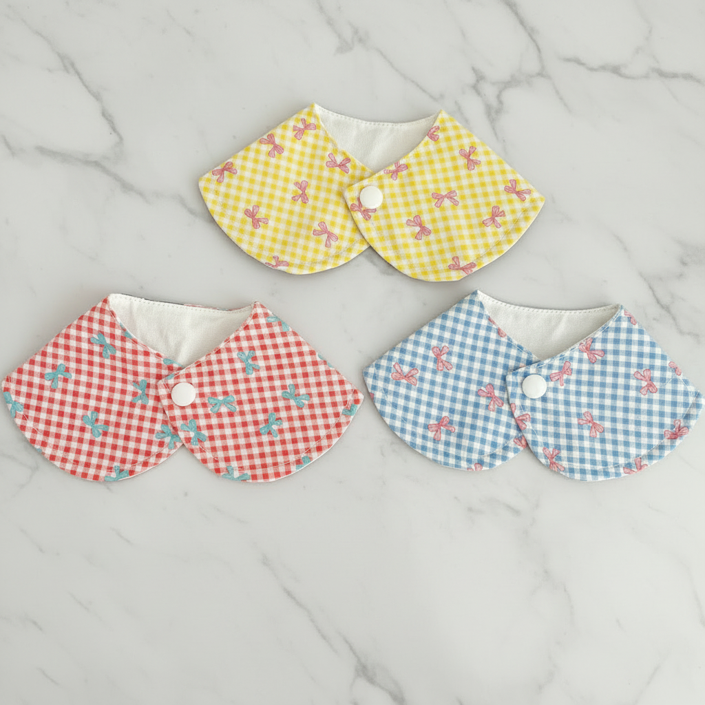 Three gingham bow pet bandanas in yellow, red, and blue on white marble background